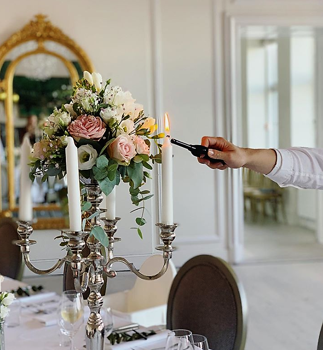 Fem armet kandelaber med blomsterdekoration i lyse duche farver. Et lys tændes på foto. Omgivelserne er på Sophienberg slot.